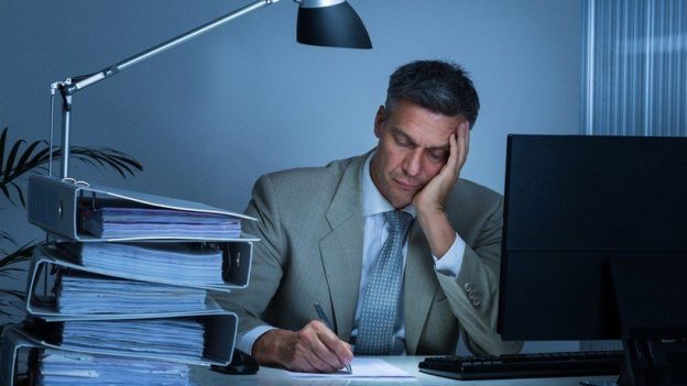 Mature man at office surrounded by full binders, lamp and computer, stressed out while working.