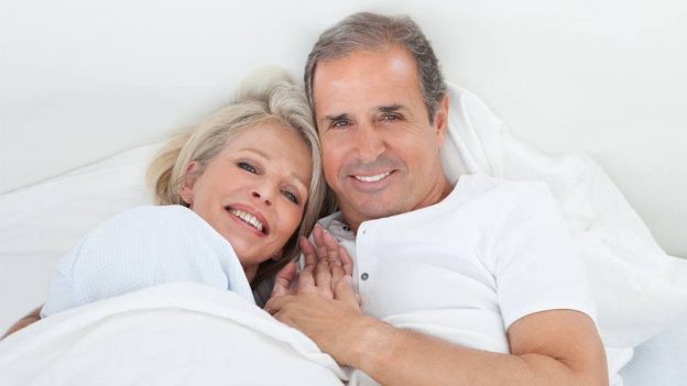 An older couple cuddled up in bed together. Low libido and sex drive can be due to testosterone deficiency.
