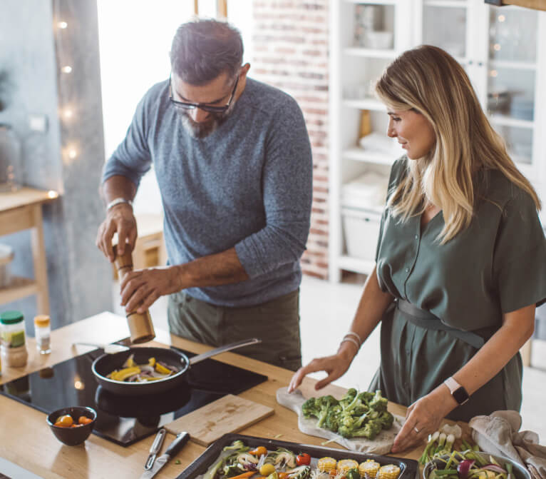Man adding salt and pepper to dish cooking on stove top as woman watches waiting to add in broccoli from chopping block, following recipes from nutrition component of program