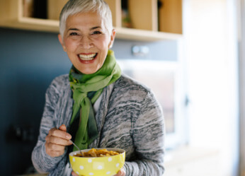 Mature woman in sweater and green scarf smiling while holding spoon and yellow bowl with nutritionally dense meal