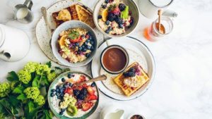 Birdseye view of a white marble table of breakfast food including waffles, oatmeal, fruit, a jar of honey, a container of cream, eggs, coffee, water, etc. Including a small bunch of green lilacs that could cause food allergies