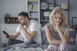 Mature man looking at phone while woman sits on opposite side of bed feeling tense experiencing anxiety and PTSD 