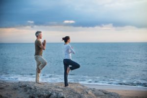 middle-aged couple doing yoga on the beach next to the water to optimize their health