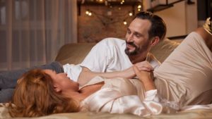 A man and woman on a bed wearing nightwear in a dimly lit room look at each other and smiling with healthy libido
