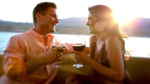 A healthy man and healthy woman sitting near a body of water smiling at each other and having a glass of wine