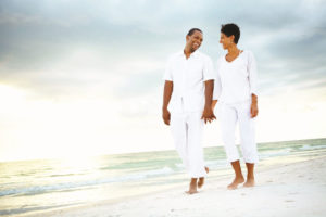 Man and woman dressed in all white holding hands and walking on the beach smiling at each other