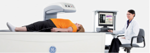 Woman in bright orange shirt laying on DEXA machine as doctor operates mechanics via computer, treating osteoporosis