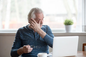 Older man with white hair holding his glasses while rubbing his eyes, man sitting at desk in office with plant in window experiencing adrenal fatigue 