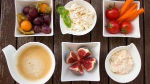 Wooden table with different foods, vegetables, fruit, dates, and dips, in white bowls and plates, that can cause food allergies 