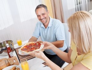Blonde woman handing pizza to a smiling mature man at dinner table, needing to get back on track with his diet