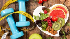 wooden table with light blue hand weights, a tape measure, cherry tomatoes, grapefruit, almonds, an apple, and a kiwi with some wheat crackers and salad, some on the table and some on a heart-shaped white plate, representing the proper diet and exercise for optimal health