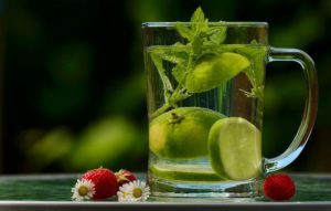 a clear mug of water with lime and a mint sprig in it with small daisies and strawberries sitting on the table next to it promoting daily water intake