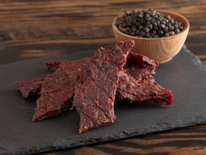 A healthy snack of beef jerky on black stone board with peppercorns in background 