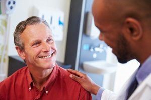 Mature male in red shirt smiling up at smiling doctor who has his hand on the man’s shoulder treating erectile dysfunction