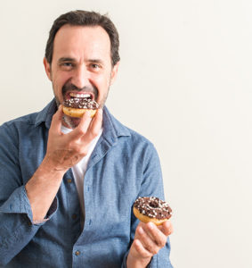 Mature man eating donut while holding a second donut in his hand that can cause high cholesterol 