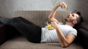 middle aged man lying on a couch eating chips/junk food that can cause visceral fat