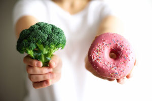 A faceless woman in a white t-shirt holding up broccoli with one hand and a donut with pink icing and sprinkles in the other that can cause high levels of LDL cholesterol