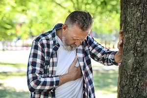 Mature man leaning against treat in park while gripping chest and trying to breathe experiencing symptoms of heart disease