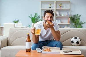 Middle-aged man eating pizza and drinking beer while sitting on a couch, not exercising and having a healthy diet that can lead to heart disease