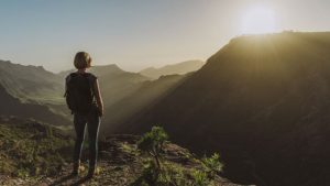 a healthy woman standing on top of a mountain with her back to the camera looking out over the valley as the sun shines from the mountain on the other side of the valley, exercising for optimal health