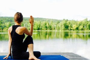 A woman seen from behind in black exercise clothes doing yoga next to a lake on a sunny day to manage stress to help lower her cholesterol levels