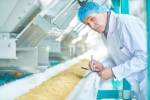 Man in food lab white coat and hair net writing on a clipboard looking at processed food