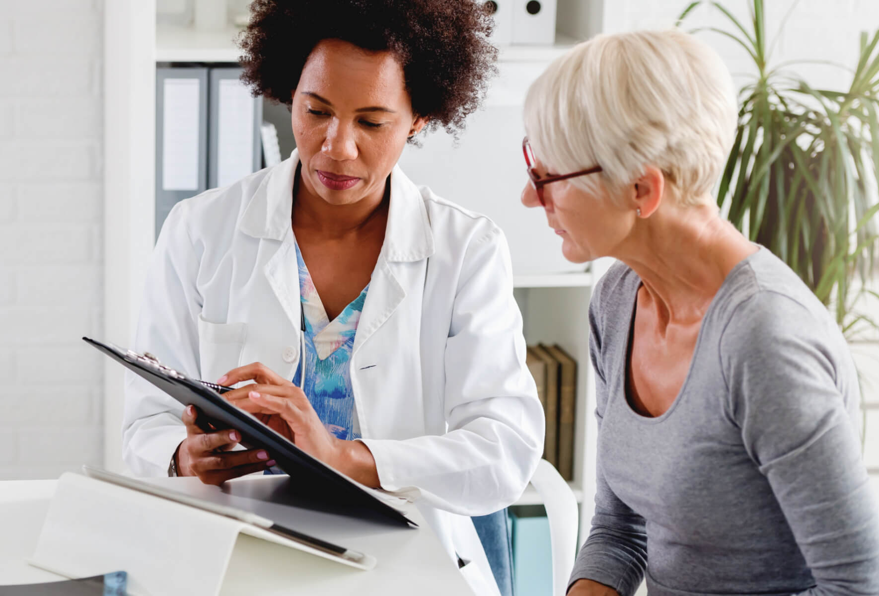 A doctor in a white coat discusses a medical form with an older woman wearing glasses and a gray shirt in a bright office setting.