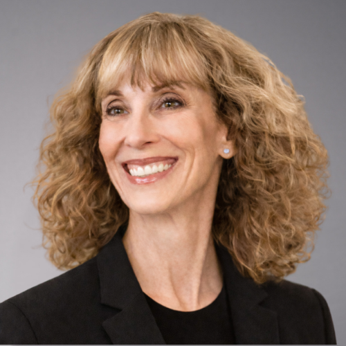 A woman with curly blonde hair and bangs smiles broadly, wearing a black blazer over a black top and a small pearl earring.