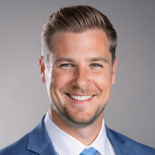 A man with short brown hair and blue eyes smiling, wearing a blue suit and light blue tie.
