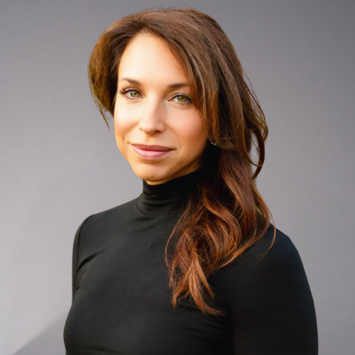 A woman with long, wavy brown hair and green eyes wears a black turtleneck and smiles slightly.