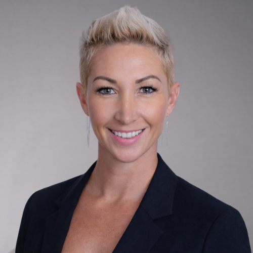 A person with short, blonde hair and green eyes wearing a black blazer and long silver earrings and smiling.