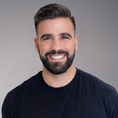 A man with dark hair and a beard smiles widely, wearing a black t-shirt against a white background.