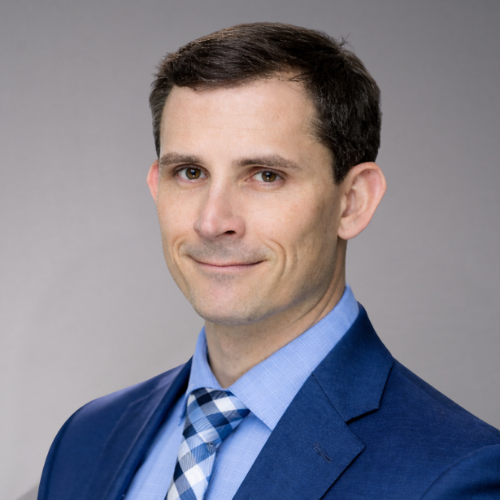 A man in a blue suit and a blue and white checkered tie smiles directly at the camera.