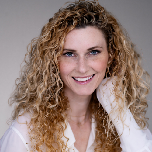 A woman with curly blonde hair and blue eyes smiles, resting her hand near her face. She is wearing a white shirt.