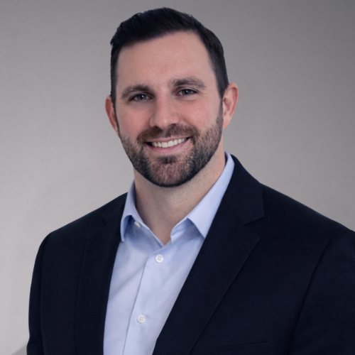 A man with short dark hair and a beard smiles at the camera. He is wearing a blue collared shirt and a dark suit jacket.