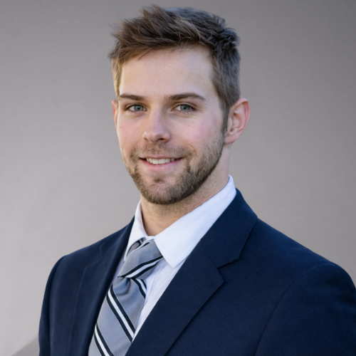 A man wearing a dark blue suit, a white dress shirt, and a gray striped tie smiles at the camera.