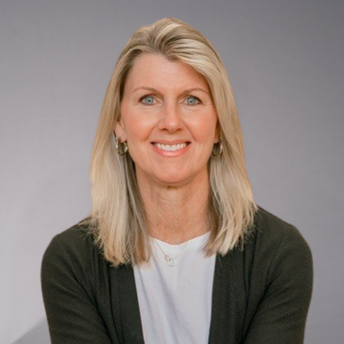 A woman with blonde hair smiles, wearing a dark cardigan over a white shirt and a silver necklace.