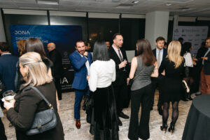 A group of people in formal attire mingling at an event, with a large banner displaying "JONA DISCOVER THE OCEAN" in the background.