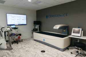 A room with medical equipment including a body composition scanner, exercise machines, and monitors displaying graphs. The Cenegenics logo is visible on the wall.