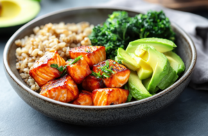 A bowl of food containing cooked salmon pieces, brown rice, sliced avocado, and green leafy vegetables.