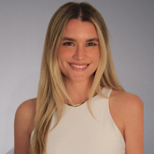 A person with long blonde hair smiles, wearing a white top and a thin gold necklace.