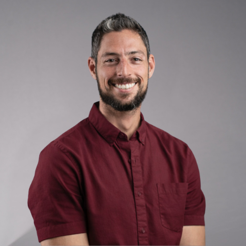 A man with short, dark hair, a beard, and a white collared shirt smiles at the camera.