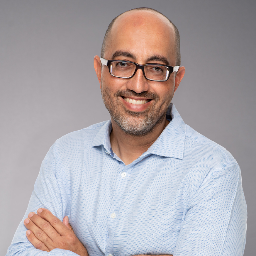 A man with glasses and a light blue patterned shirt smiles with his arms crossed.