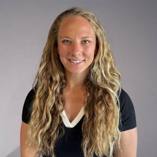 A person with long, curly blonde hair smiles at the camera. They are wearing a black t-shirt with a white V-neck accent.